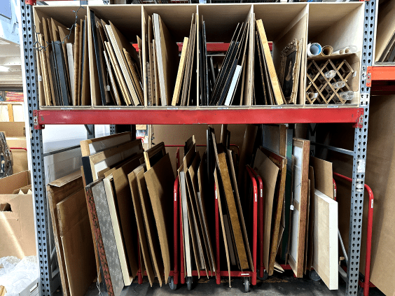 Framed artworks stacked on warehouse shelving from an estate collection